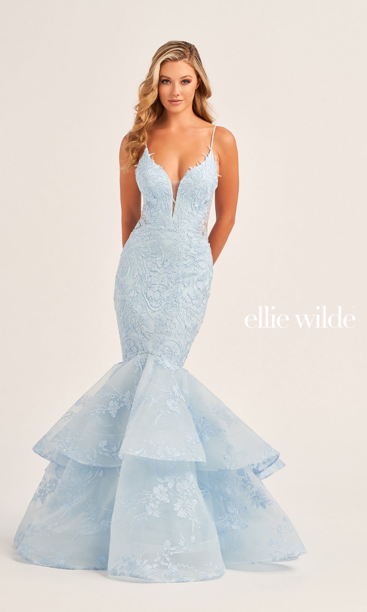 A woman stands against a plain background in the Ellie Wilde Long Lace Mermaid Prom Dress EW35038, featuring light blue beaded lace, thin straps, a deep V neckline, and a layered flared mermaid skirt.